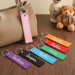 Op de afbeelding: Een verzameling gepersonaliseerde sleutelhangers in verschillende kleuren, waaronder roze, oranje, rood, groen, blauw, paars en zwart. Elke sleutelhanger heeft een naam in wit script geborduurd. Eén roze sleutelhanger is bevestigd aan een beige handtas.