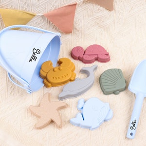 Peut inclure: Un seau en plastique bleu avec le nom "Bella" inscrit dessus, ainsi qu'un ensemble de moules à sable en silicone colorés en forme de crabe, de tortue, de dauphin, de coquille Saint-Jacques et d'étoile de mer. L'ensemble comprend également une pelle en plastique bleue avec le nom "Bella" inscrit dessus.