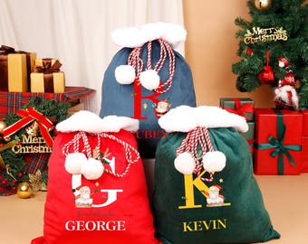May include: Three plush Christmas gift bags in red, blue, and green. Each bag has a white fluffy trim, a drawstring with pom-poms, and a large initial with a Santa Claus graphic. The names "GEORGE", "RUBEN", and "KEVIN" are printed on the bags.