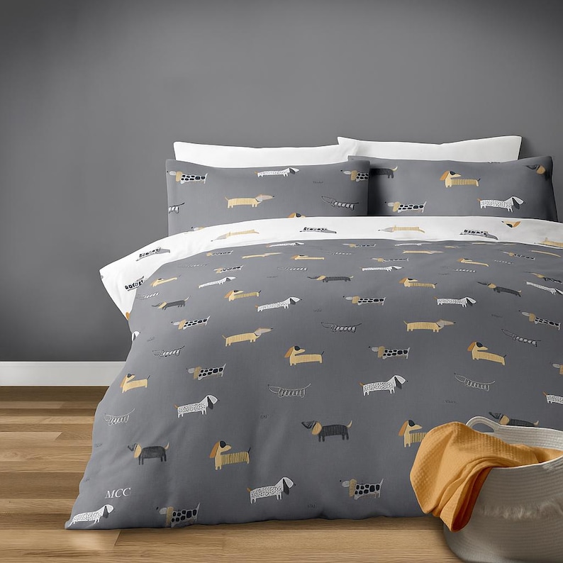 May include: Gray duvet cover and pillowcases set with a repeating pattern of dachshund dogs in various colors. The bedding set includes a duvet cover and two pillowcases. The duvet cover has a white underside. A basket with an orange blanket is in the corner.