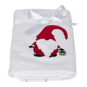 May include: White bath towel with an embroidered gnome holding a lantern. The gnome is red and white. The towel is tied with a white ribbon.