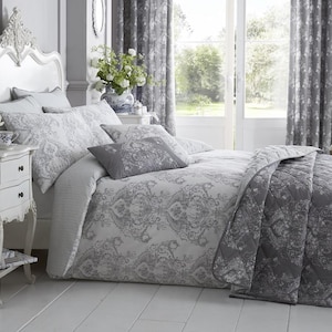 Pode incluir: Um quarto com uma cama vestida com roupa de cama floral cinza e branca, incluindo uma capa de edredão, almofadas e um edredão acolchoado. Uma cabeceira e uma mesa de cabeceira brancas são visíveis, juntamente com cortinas cinzentas estampadas e uma lâmpada.
