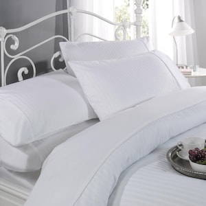 May include: White bed linens with a subtle stripe pattern. The bed is made with a white duvet cover, two white pillowcases, and a white bed skirt. The bed is set against a gray wall and has a white metal headboard.