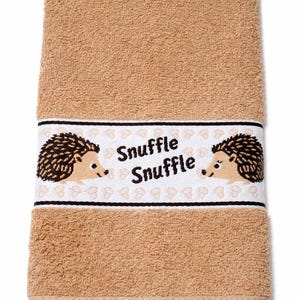 Könnte beinhalten: Beige Badetuch mit einem dekorativen Band, das zwei Cartoon-Igel und den Text "Snuffle Snuffle" zeigt. Das Handtuch ist rechteckig und aus einem weichen, saugfähigen Material gefertigt.