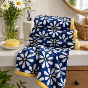 Pode incluir: Uma pilha de três toalhas azul-marinho dobradas e uma toalha estendida, todas com um padrão repetitivo de margaridas brancas com centros amarelos. Cada toalha é finalizada com uma borda amarela sólida nas extremidades, criando um design floral.
