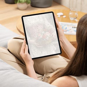 May include: A black tablet displaying a blurred, abstract image with red and white tones. The tablet is held by a person wearing tan pants, sitting on a gray beanbag chair. A small wooden table and a glass of water are in the background.