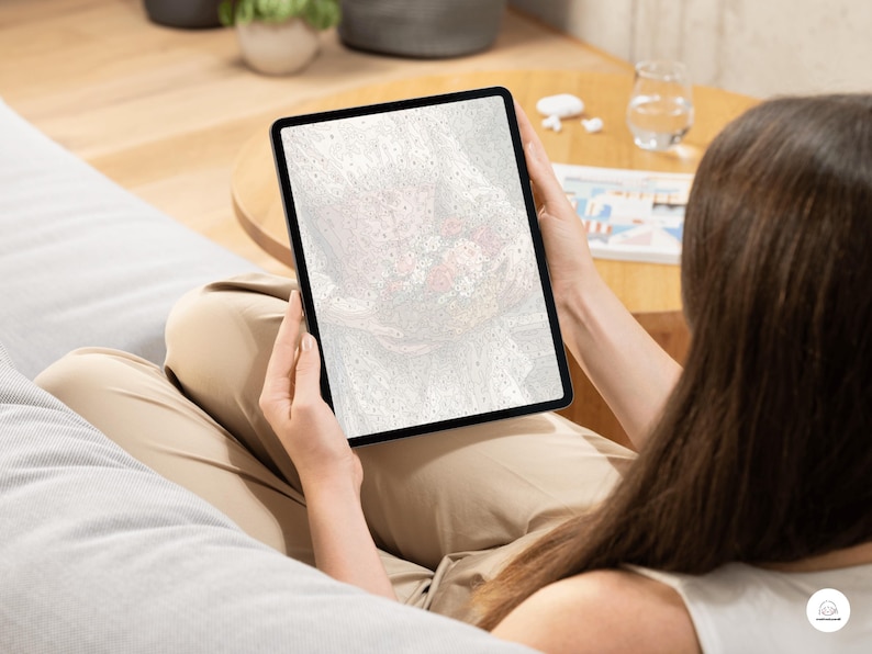 May include: A black tablet displaying a blurred, abstract image with red and white tones. The tablet is held by a person wearing tan pants, sitting on a gray beanbag chair. A small wooden table and a glass of water are in the background.