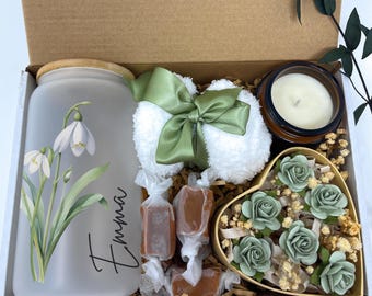 May include: A gift box filled with a variety of items, including a frosted glass jar with a floral design and the name "Emma" written on it, a white towel with a green bow, a brown glass candle, a heart-shaped box filled with green paper flowers, and a bag of caramel candies.
