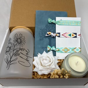 May include: A gift box containing a frosted glass jar with a floral design and the name "Chloe", a teal journal, hair ties, a white rose, and a candle. The items are arranged on decorative straw.