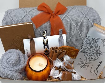 May include: A gift box containing a gray textured blanket with an orange ribbon, a brown notebook, a frosted glass tumbler with floral art and the name Emma, a pumpkin-shaped candle, cinnamon sticks, dried orange slices, and hair ties.