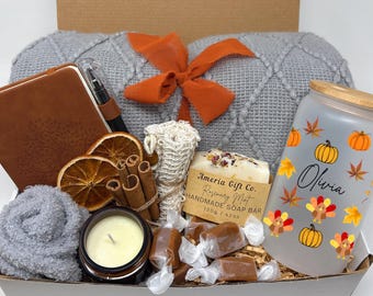 May include: A gift box containing a gray knit blanket with an orange ribbon, a brown notebook with a pen, dried orange slices, cinnamon sticks, a candle, a loofah, a soap bar labeled Ameria Gift Co. Rosemary Mint 120g / 4.2oz, caramels, and a glass tumbler with a bamboo lid featuring the name Olivia, pumpkins, turkeys, and autumn leaves.