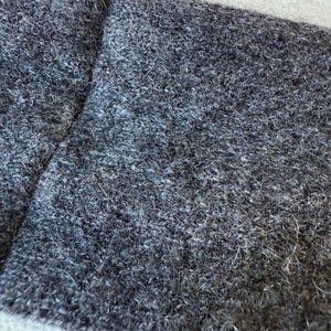 May include: Close-up of a grey, textured fabric with a handmade tag. The fabric appears to be a soft, woven material, possibly wool or a wool blend. The tag has the word "HANDMADE" in a script font.