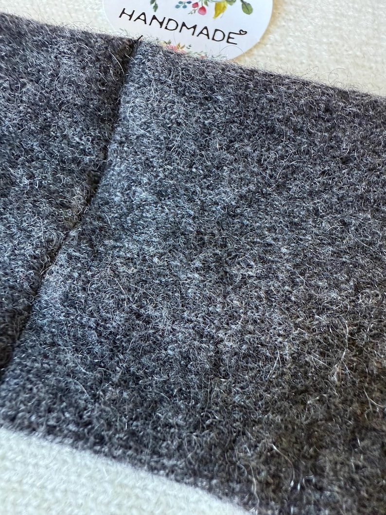 May include: Close-up of a grey, textured fabric with a handmade tag. The fabric appears to be a soft, woven material, possibly wool or a wool blend. The tag has the word "HANDMADE" in a script font.