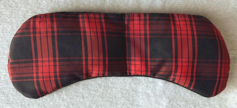 May include: A red and black plaid eye mask. The mask is rectangular with rounded edges and a curved bottom. The plaid pattern features thick red and black lines. The mask is designed to block out light.