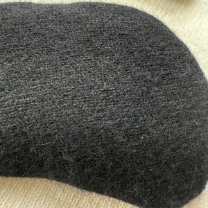 May include: Close-up of a dark gray, oval-shaped knitted item, possibly a hat or a part of a garment. The texture appears soft and fuzzy. A portion of a black and red striped object is visible in the background.
