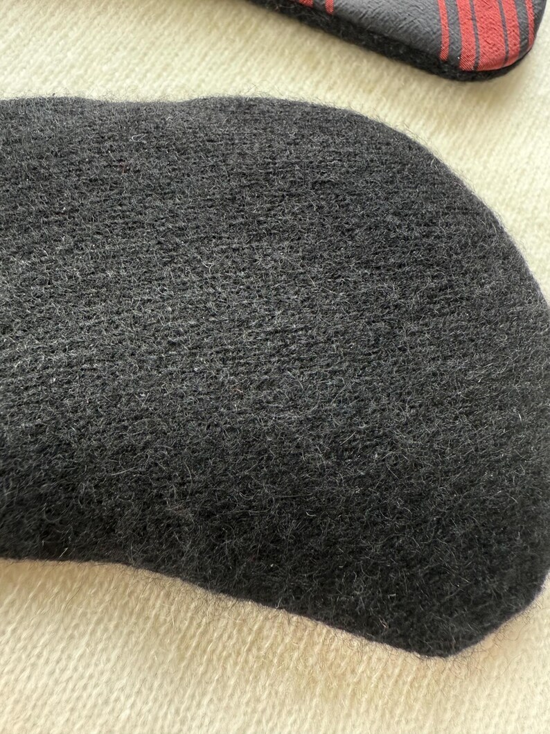 May include: Close-up of a dark gray, oval-shaped knitted item, possibly a hat or a part of a garment. The texture appears soft and fuzzy. A portion of a black and red striped object is visible in the background.