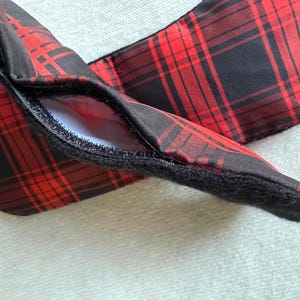 May include: A red and black plaid fabric item with a hidden pocket, featuring a black zipper and a soft, fuzzy edge. The plaid pattern is a classic design, and the item appears to be a scarf or similar accessory.