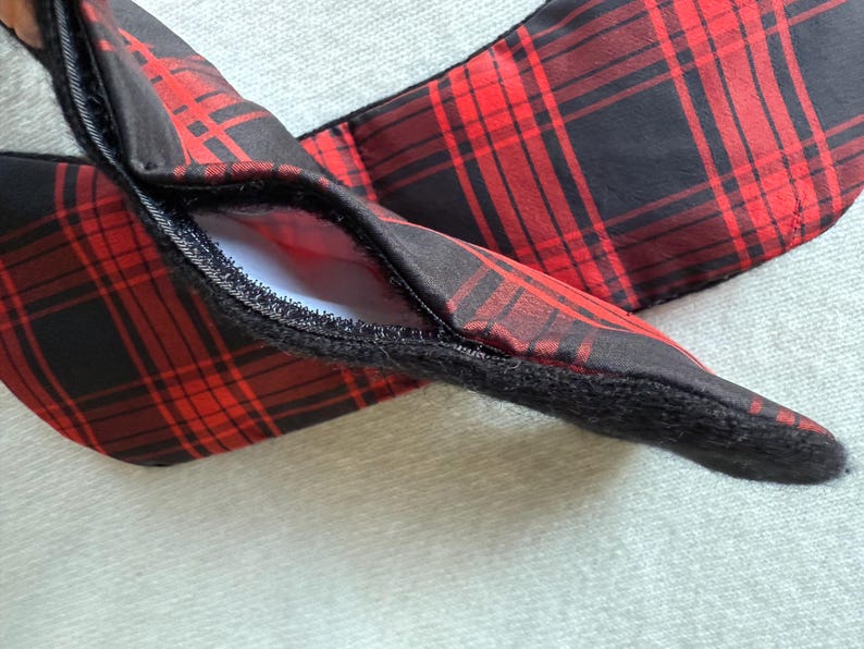 May include: A red and black plaid fabric item with a hidden pocket, featuring a black zipper and a soft, fuzzy edge. The plaid pattern is a classic design, and the item appears to be a scarf or similar accessory.