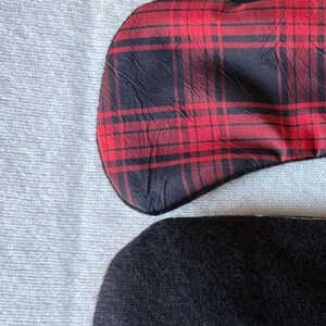May include: Two sleep masks. One is red and black plaid, the other is solid black. Both are curved at the top and bottom, and are resting on a white, textured surface.