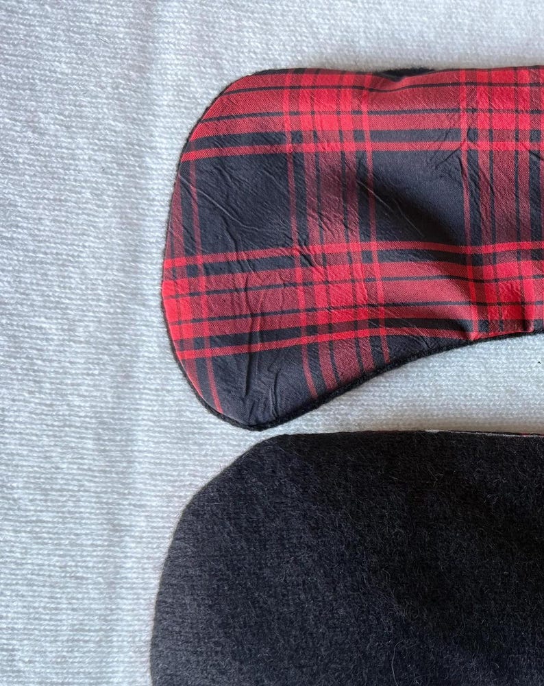 May include: Two sleep masks. One is red and black plaid, the other is solid black. Both are curved at the top and bottom, and are resting on a white, textured surface.