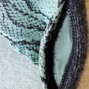 May include: Close-up of a fabric with a teal and black pattern. The fabric has a textured appearance and is folded to reveal a contrasting interior. A dark, fuzzy trim borders the edge of the fabric.