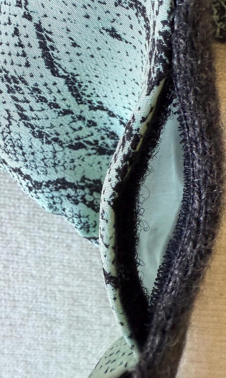 May include: Close-up of a fabric with a teal and black pattern. The fabric has a textured appearance and is folded to reveal a contrasting interior. A dark, fuzzy trim borders the edge of the fabric.