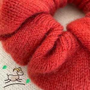 May include: A close-up of a red knitted hair scrunchie. The scrunchie is made of a soft, fuzzy material and has a textured surface.