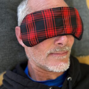 May include: A red and black plaid eye mask covers the eyes of a person. The mask is made of fabric and has a rectangular shape. The person is wearing a black hooded sweatshirt.