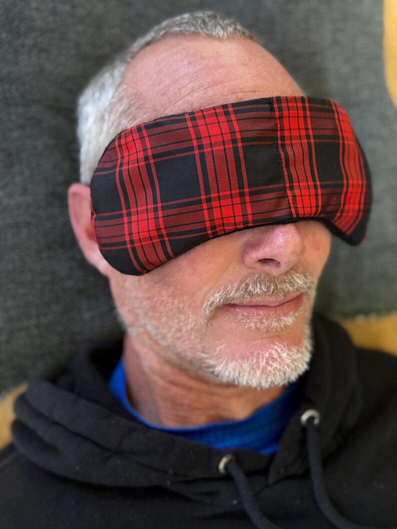 May include: A red and black plaid eye mask covers the eyes of a person. The mask is made of fabric and has a rectangular shape. The person is wearing a black hooded sweatshirt.