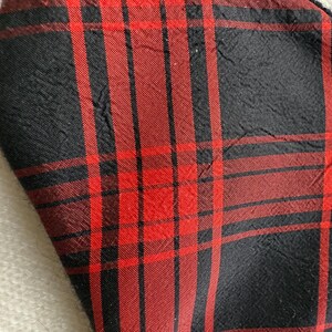 May include: Close-up of a fabric with a red and black plaid pattern. The fabric has a textured appearance with visible creases. The plaid design features vertical and horizontal lines creating a grid-like structure. The color scheme is dominated by red and black.