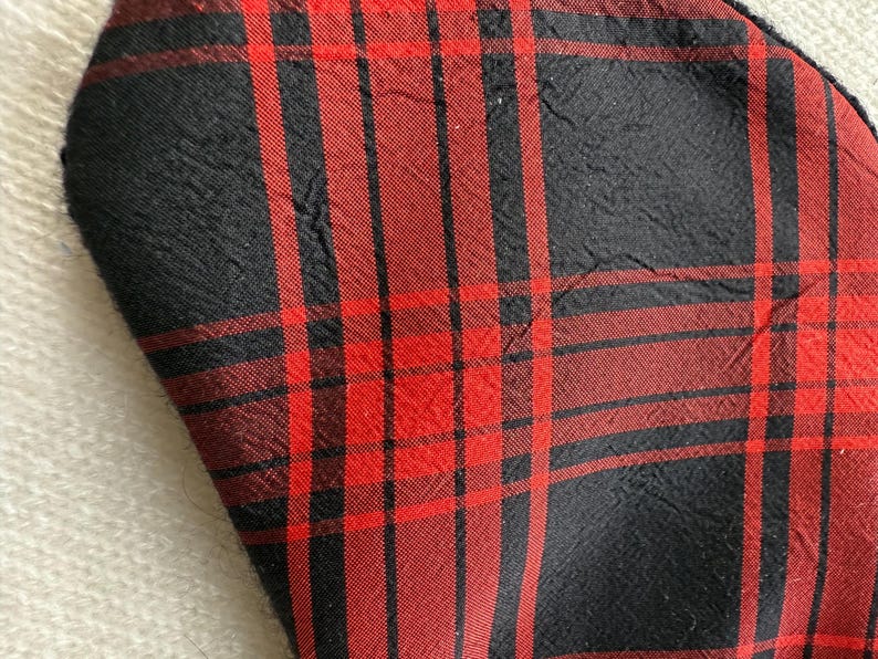 May include: Close-up of a fabric with a red and black plaid pattern. The fabric has a textured appearance with visible creases. The plaid design features vertical and horizontal lines creating a grid-like structure. The color scheme is dominated by red and black.