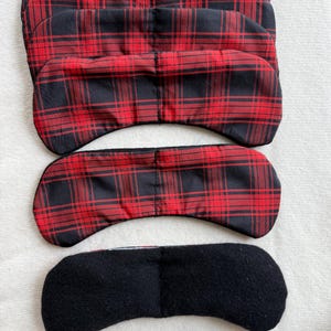 May include: Several eye masks in a plaid pattern of red and black are displayed. One mask is black. The masks are made of fabric and are designed to block out light. The masks are designed to be worn over the eyes.