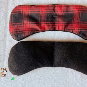 May include: Two eye masks. The top mask has a red and black plaid pattern. The bottom mask is solid black. Both masks are curved to fit the eye area. The masks are on a white surface.