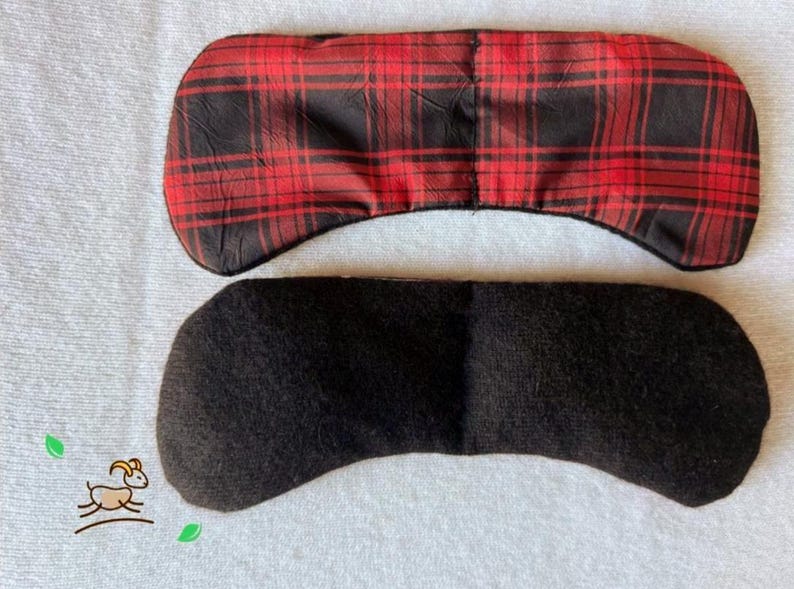May include: Two eye masks. The top mask has a red and black plaid pattern. The bottom mask is solid black. Both masks are curved to fit the eye area. The masks are on a white surface.