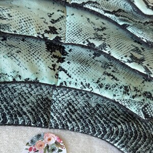 May include: Close-up of a handmade item with a snakeskin pattern in teal and black. The item is layered and textured. A round sticker with the word "HANDMADE" and floral accents is visible.