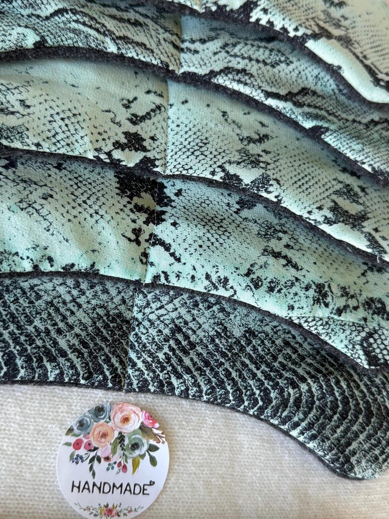 May include: Close-up of a handmade item with a snakeskin pattern in teal and black. The item is layered and textured. A round sticker with the word "HANDMADE" and floral accents is visible.