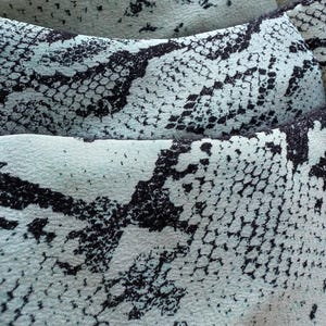 May include: Close-up of a fabric with a snakeskin pattern. The material features a pale blue background with black, irregular shapes resembling snake scales. The texture appears soft and slightly wrinkled, suggesting a flowing material.