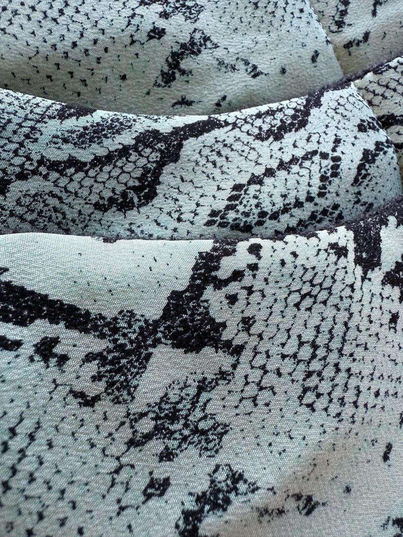 May include: Close-up of a fabric with a snakeskin pattern. The material features a pale blue background with black, irregular shapes resembling snake scales. The texture appears soft and slightly wrinkled, suggesting a flowing material.
