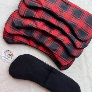 May include: A stack of five reusable menstrual pads with a red and black plaid pattern. One black pad is displayed separately, showing the underside. A small round sticker with the word "handmade" is also visible.