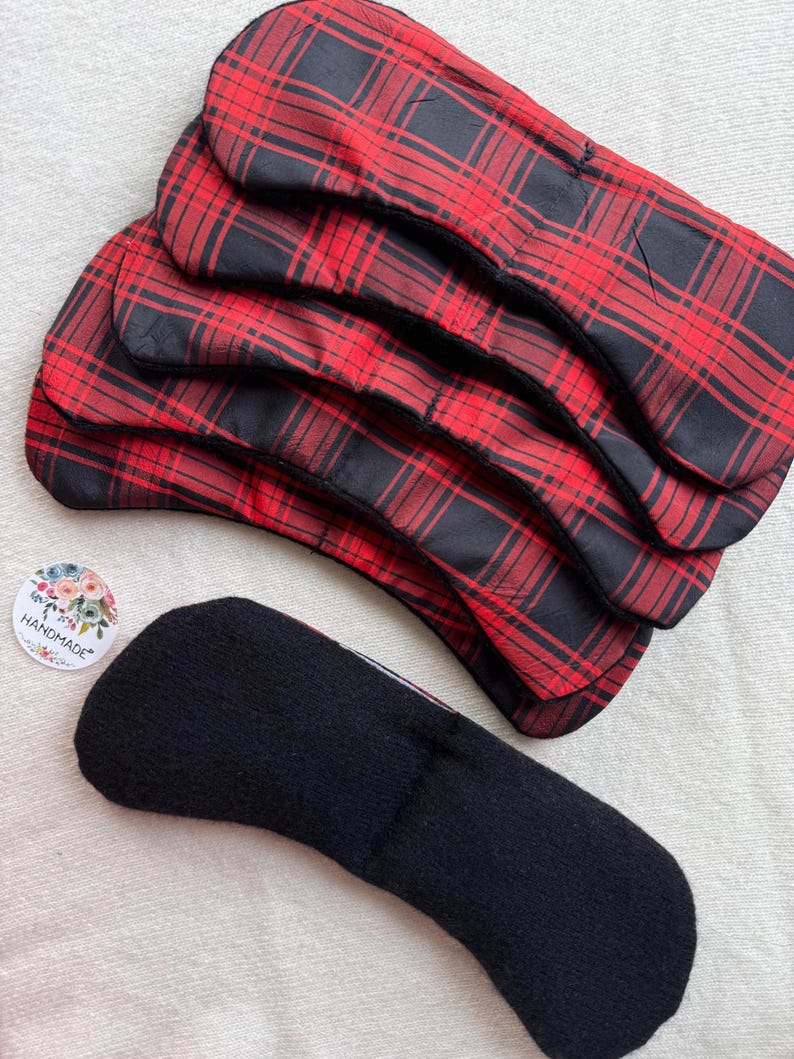 May include: A stack of five reusable menstrual pads with a red and black plaid pattern. One black pad is displayed separately, showing the underside. A small round sticker with the word "handmade" is also visible.