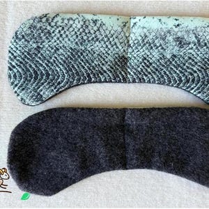May include: Two eye masks. The top mask has a snake skin pattern in shades of teal and black. The bottom mask is a solid dark grey. Both masks are curved and made of fabric.