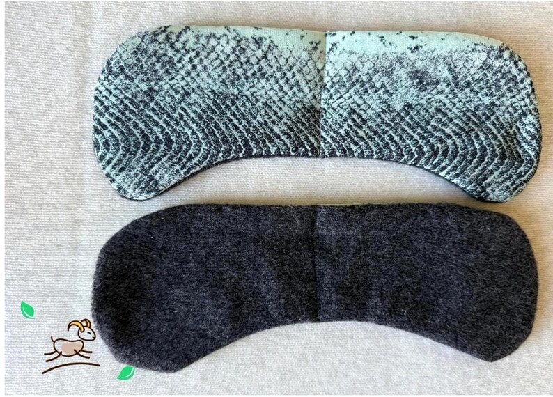 May include: Two eye masks. The top mask has a snake skin pattern in shades of teal and black. The bottom mask is a solid dark grey. Both masks are curved and made of fabric.
