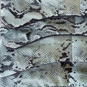 May include: Close-up of a fabric with a snakeskin pattern. The fabric is a light teal colour with black markings, creating a reptile-like texture. The image shows multiple layers of the fabric, highlighting its design and texture.