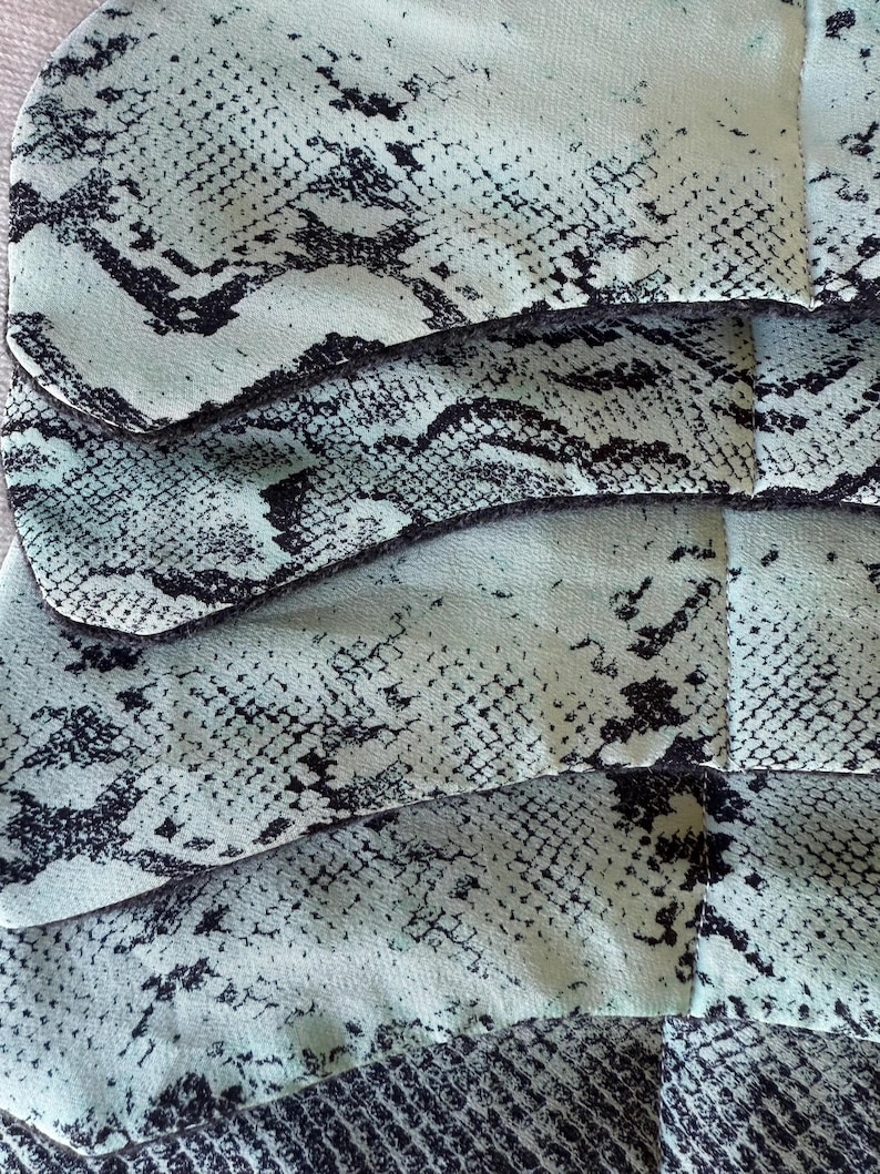 May include: Close-up of a fabric with a snakeskin pattern. The fabric is a light teal colour with black markings, creating a reptile-like texture. The image shows multiple layers of the fabric, highlighting its design and texture.