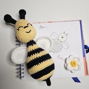 May include: A handmade crochet bee with black and yellow stripes, white wings, and a smiling face. The bee is placed next to a drawing of a bee and a white crochet flower with a yellow center. The word "Bee" and the name "Beatrice" are written on the page.
