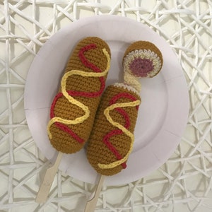 Peut inclure: Deux corndogs en crochet sur des bâtonnets blancs, avec de la moutarde jaune et du ketchup rouge, sur une assiette blanche. Les corndogs sont bruns, jaunes et rouges.