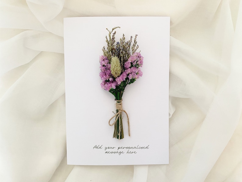 Op de afbeelding: Een witte kaart met een klein boeket gedroogde bloemen, waaronder paarse en witte bloesems, en een bruine touw strik. De kaart heeft de tekst "Voeg uw persoonlijke boodschap hier toe" erop geschreven.