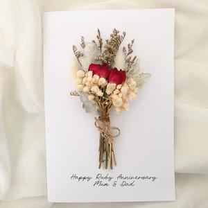 May include: A white greeting card with a bouquet of dried flowers. The bouquet includes two red roses, cream-colored baby's breath, and silver-green leaves, tied with twine. The card reads "Happy Ruby Anniversary Mum & Dad."