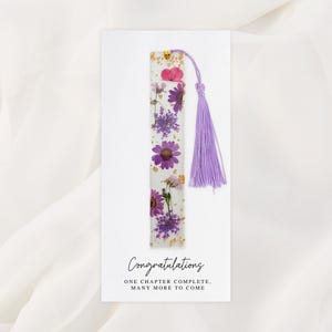 May include: A white bookmark with embedded purple and pink flowers and a lavender tassel. The bookmark is on a white card with the words "Congratulations" and "ONE CHAPTER COMPLETE, MANY MORE TO COME" in black script.