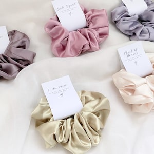 May include: Set of five satin scrunchies with different colors and text cards. The cards have different titles, including "Bride", "Bride Squad", "Maid of Honour", and "I do Crew".